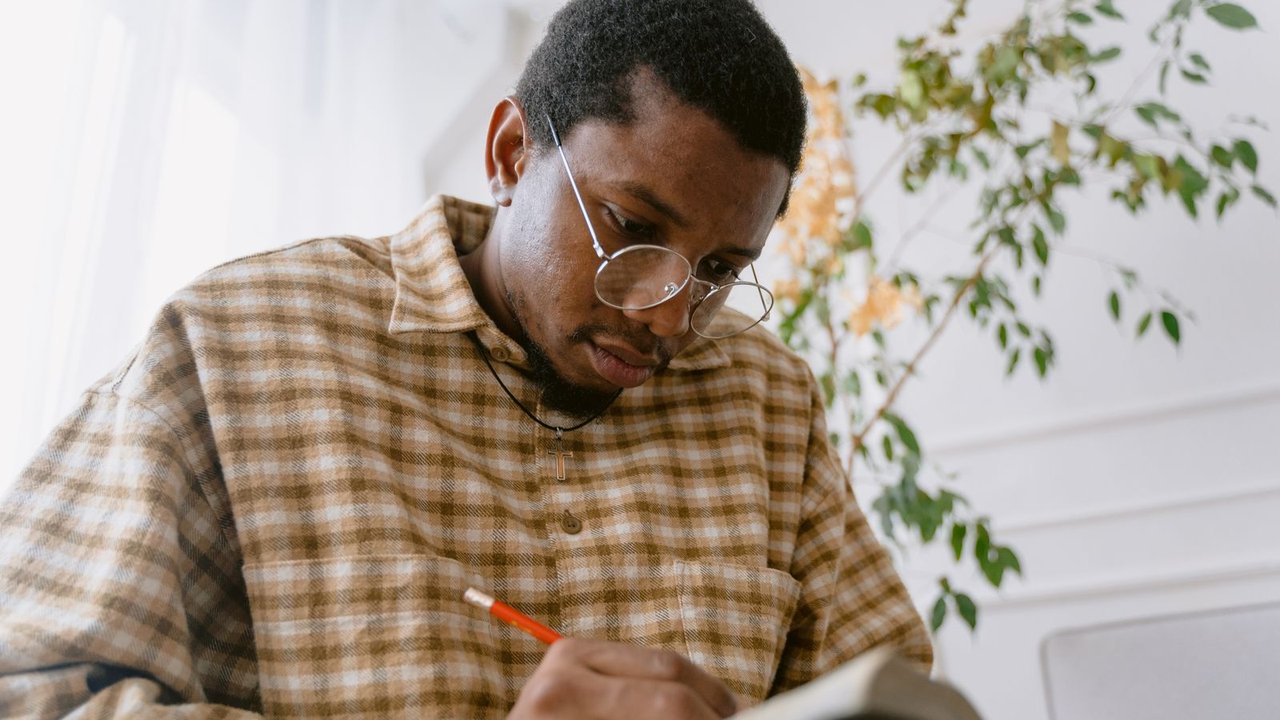 A man in glasses writing in a notebook by a window, deep in thought.