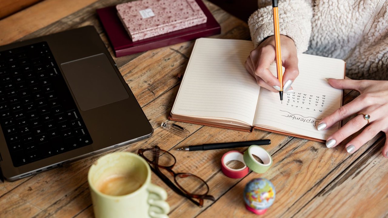 Hands writing a plan in a notebook beside a laptop and a mug of coffee on a wooden desk.