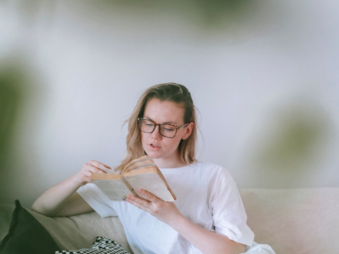 A woman relaxing on a pale sofa in soft daylight, reading a book.